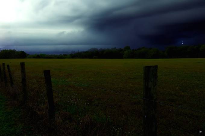 Dark storm clouds gathering over a green field with a wooden fence.